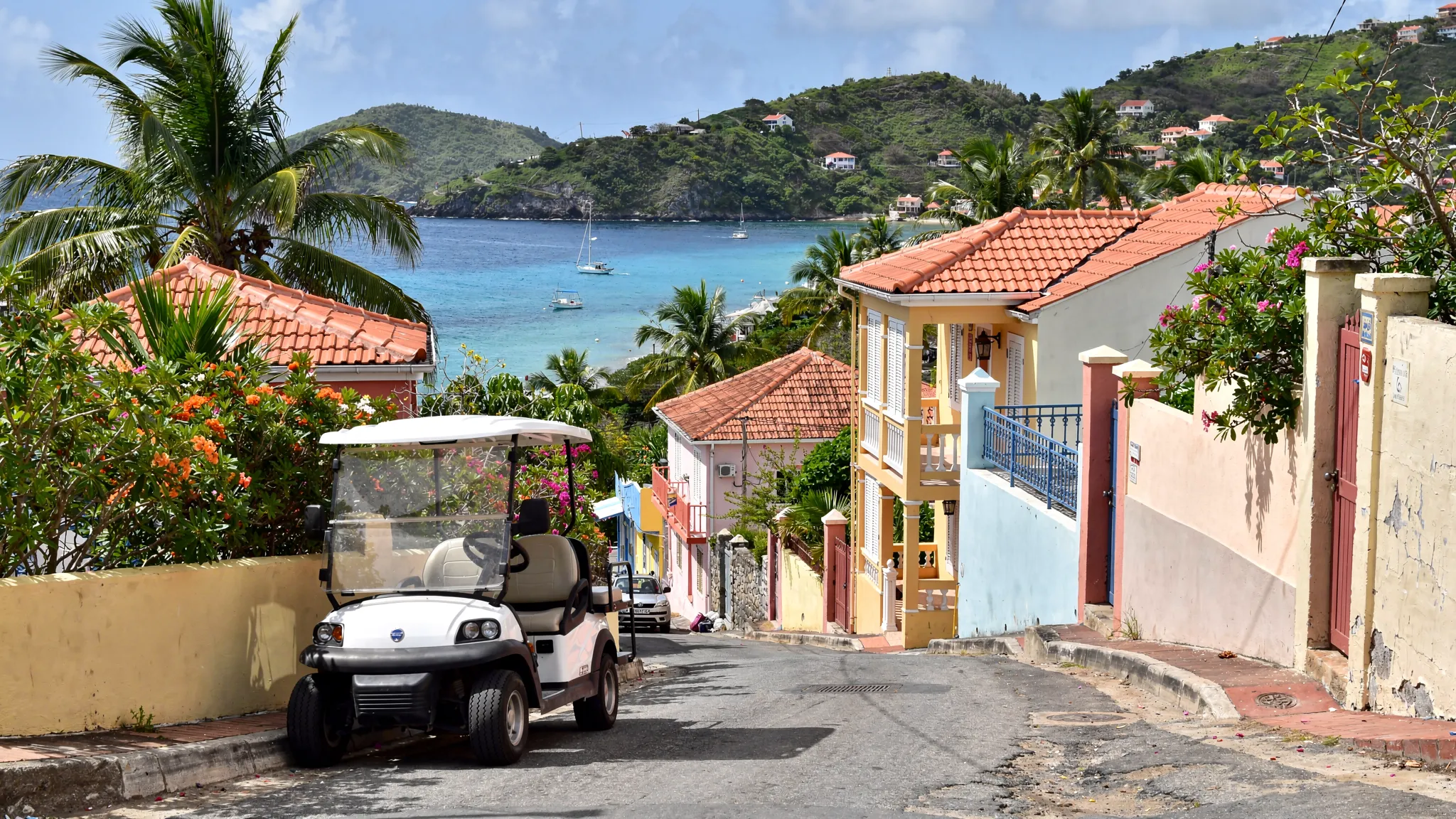 Une voiturette électrique stationnée à Terre-de-Haut, avec en arrière-plan une rue en pente typique des Saintes, de la végétation tropicale et un aperçu de la baie au loin.