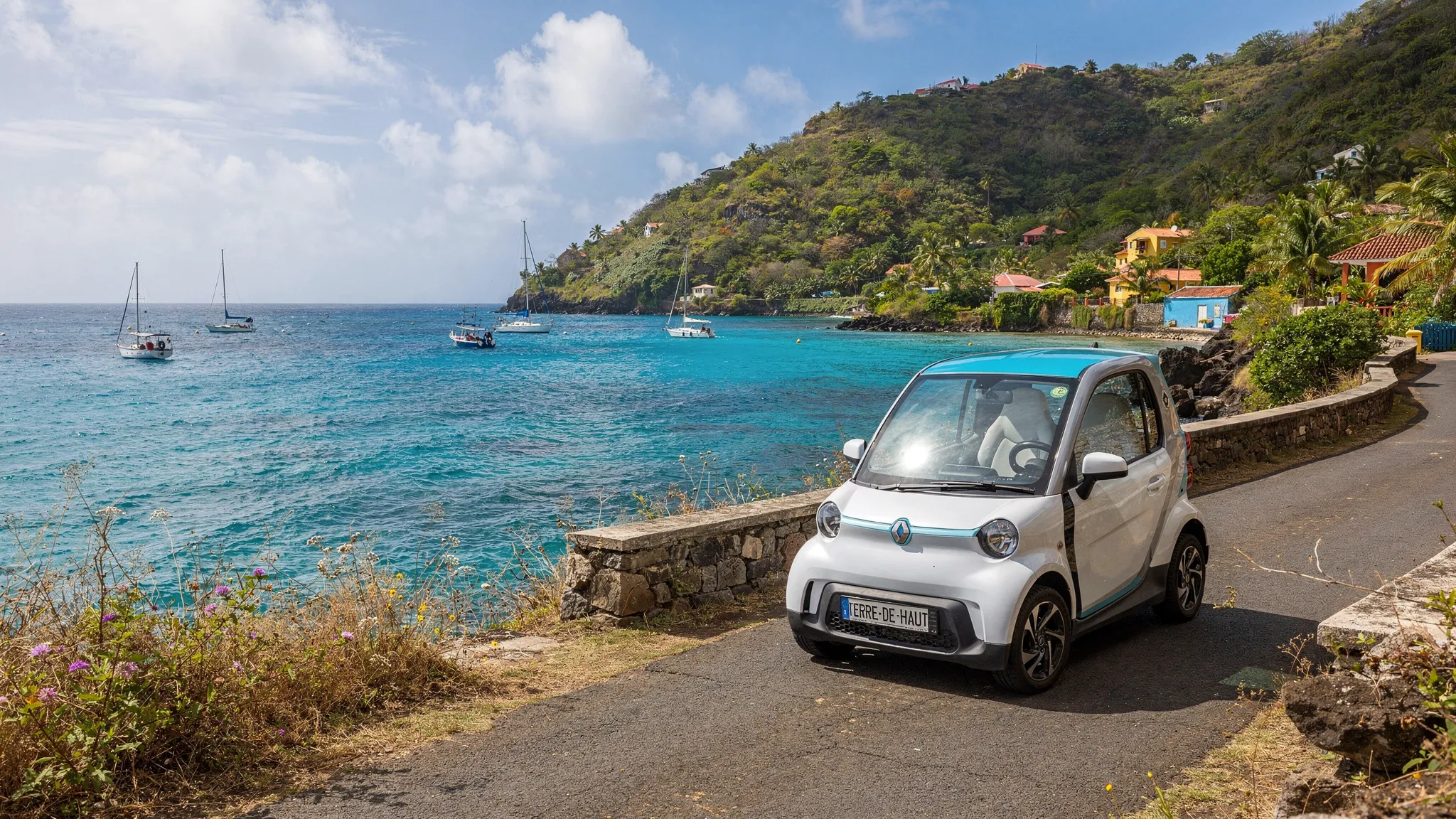 Une voiturette électrique stationnée près d’une route côtière à Terre-de-Haut, avec la mer des Saintes, des collines verdoyantes et quelques bateaux au loin.