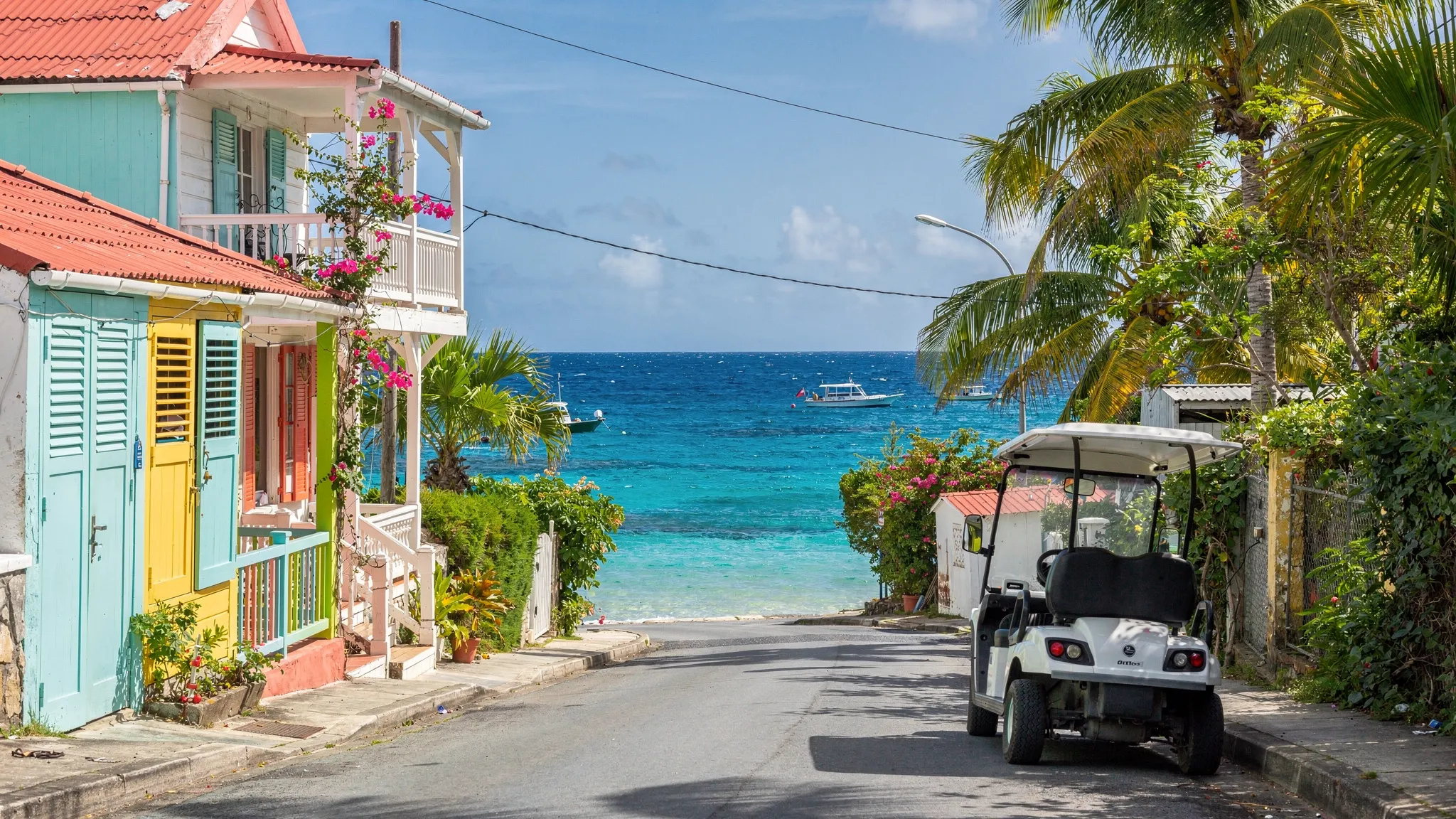 Une voiturette électrique stationnée près d'une rue calme de Terre-de-Haut, avec la mer des Saintes en arrière-plan, des maisons créoles colorées et une ambiance de journée ensoleillée.