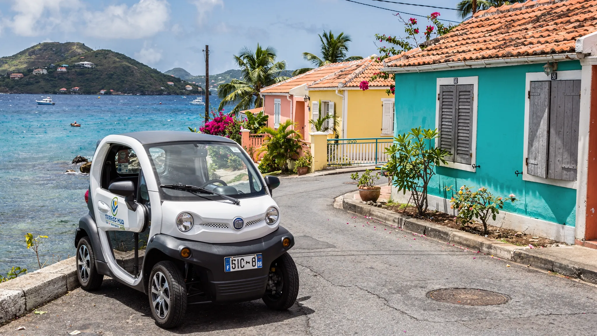 Une voiturette électrique de location stationnée à Terre-de-Haut avec en arrière-plan une route côtière, des cases colorées et la mer des Saintes.