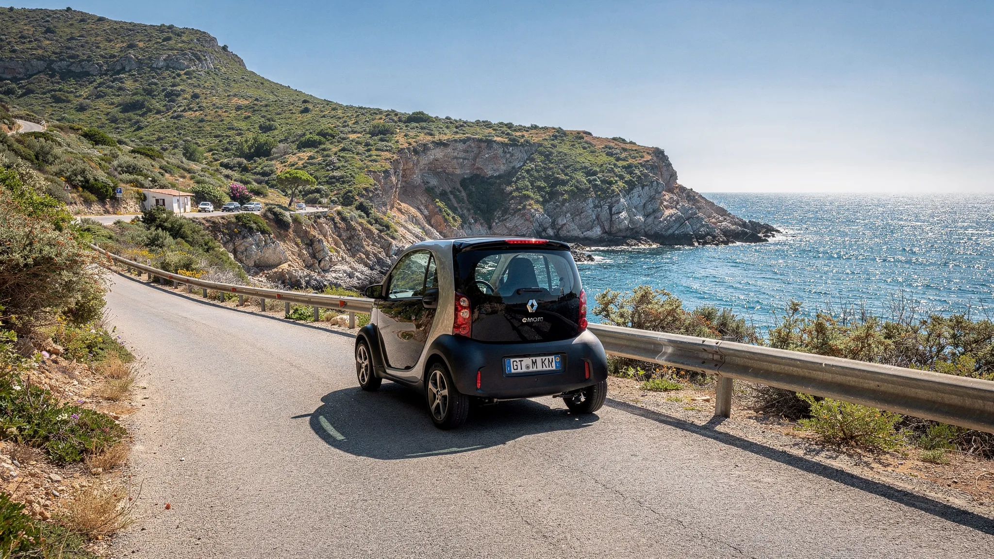 Une voiturette électrique roule sur une route étroite et ensoleillée de bord de mer, avec des collines en arrière-plan et une petite zone de stationnement au loin.