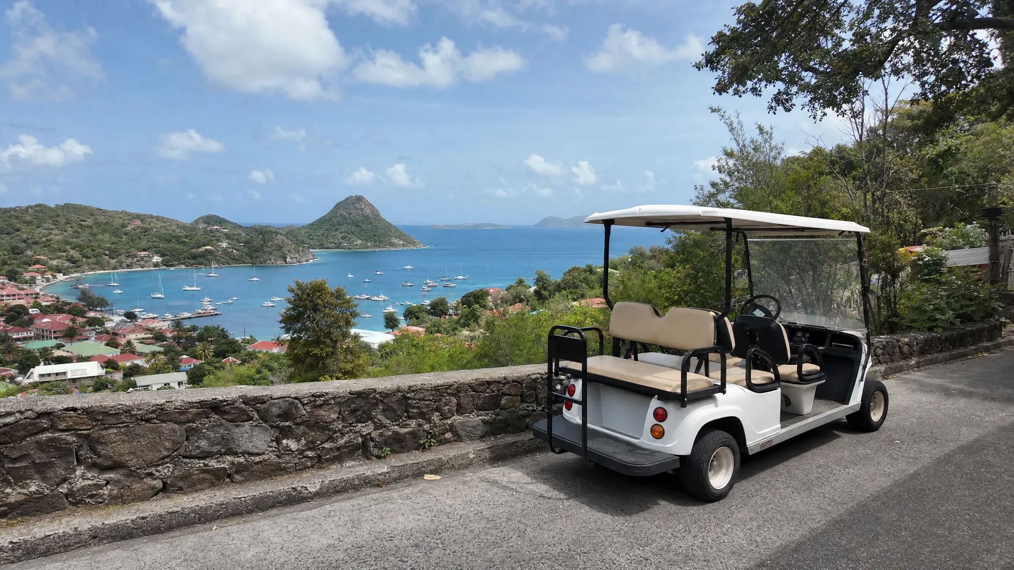Une voiturette électrique stationnée au bord d’une route étroite à Terre-de-Haut, avec un muret en pierre, la baie des Saintes en contrebas et la mer au loin.