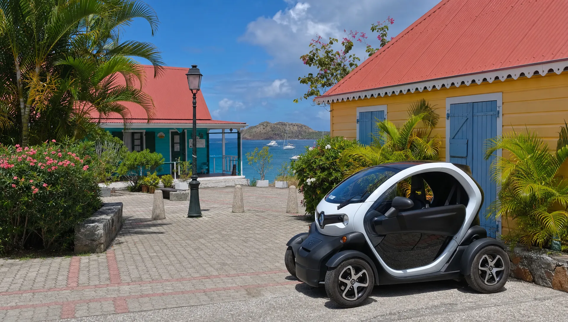 Une voiturette électrique stationnée au bord d’une petite place de Terre-de-Haut, avec des maisons créoles colorées, quelques plantes tropicales et la mer visible entre les bâtiments.